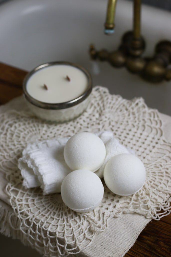 3 all natural diy bath bombs sitting next to a clawfoot bathtub with a brass faucet. They're sitting on a white washcloth and a vintage doily next to a candle in a china glass bowl with a silver rim.