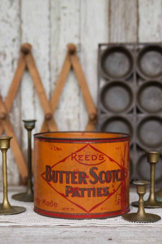 vignette of vintage brass candlesticks, a muffin pan, an orange reed's butterscotch patties tin and a coat hanger with a chippy white wood background
