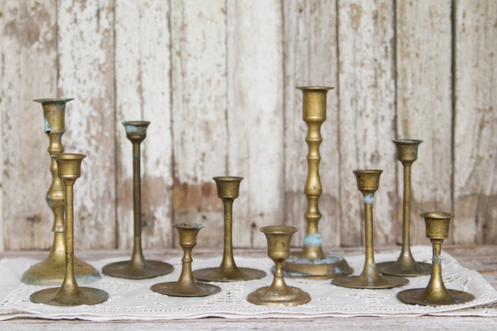 assortment of vintage brass candlesticks on an antique linen table runner with a white chippy paint wooden bacgdrop