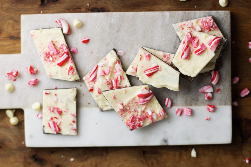 pieces of peppermint bark on a gray marble cutting board with peppermint pieces and white chocolate chips scattered around it
